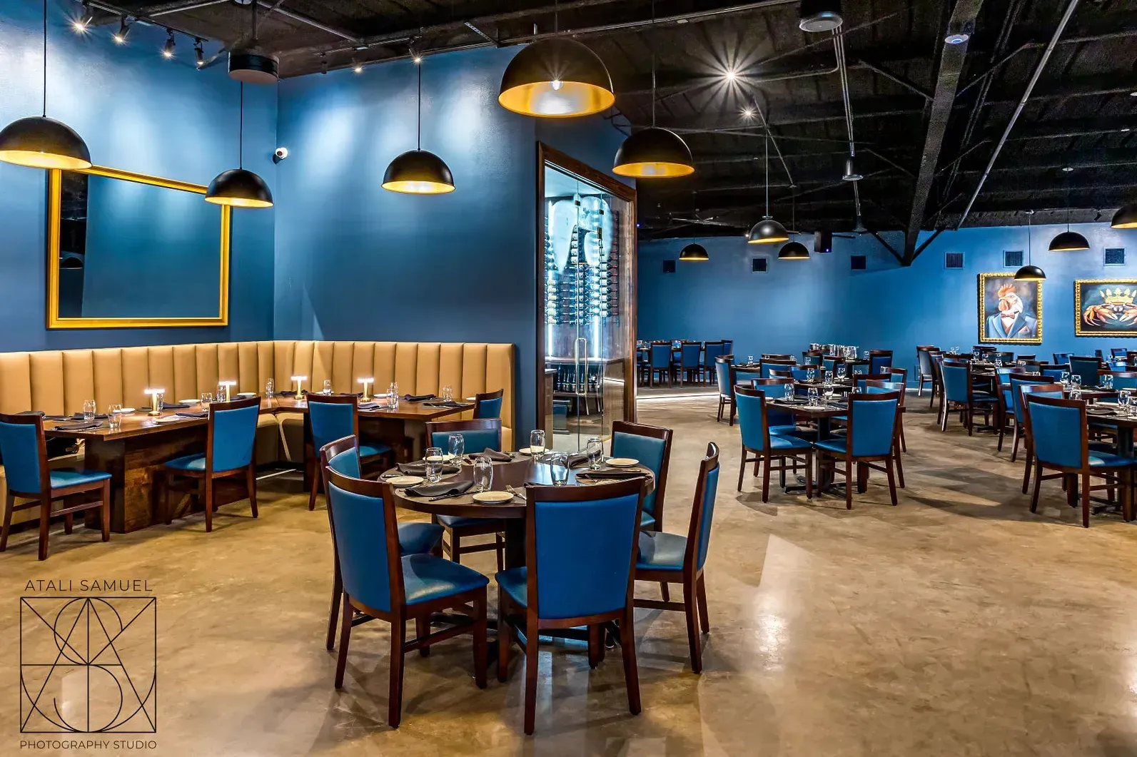 Restaurant interior with blue walls, round tables, blue chairs, and hanging black and gold lamps.