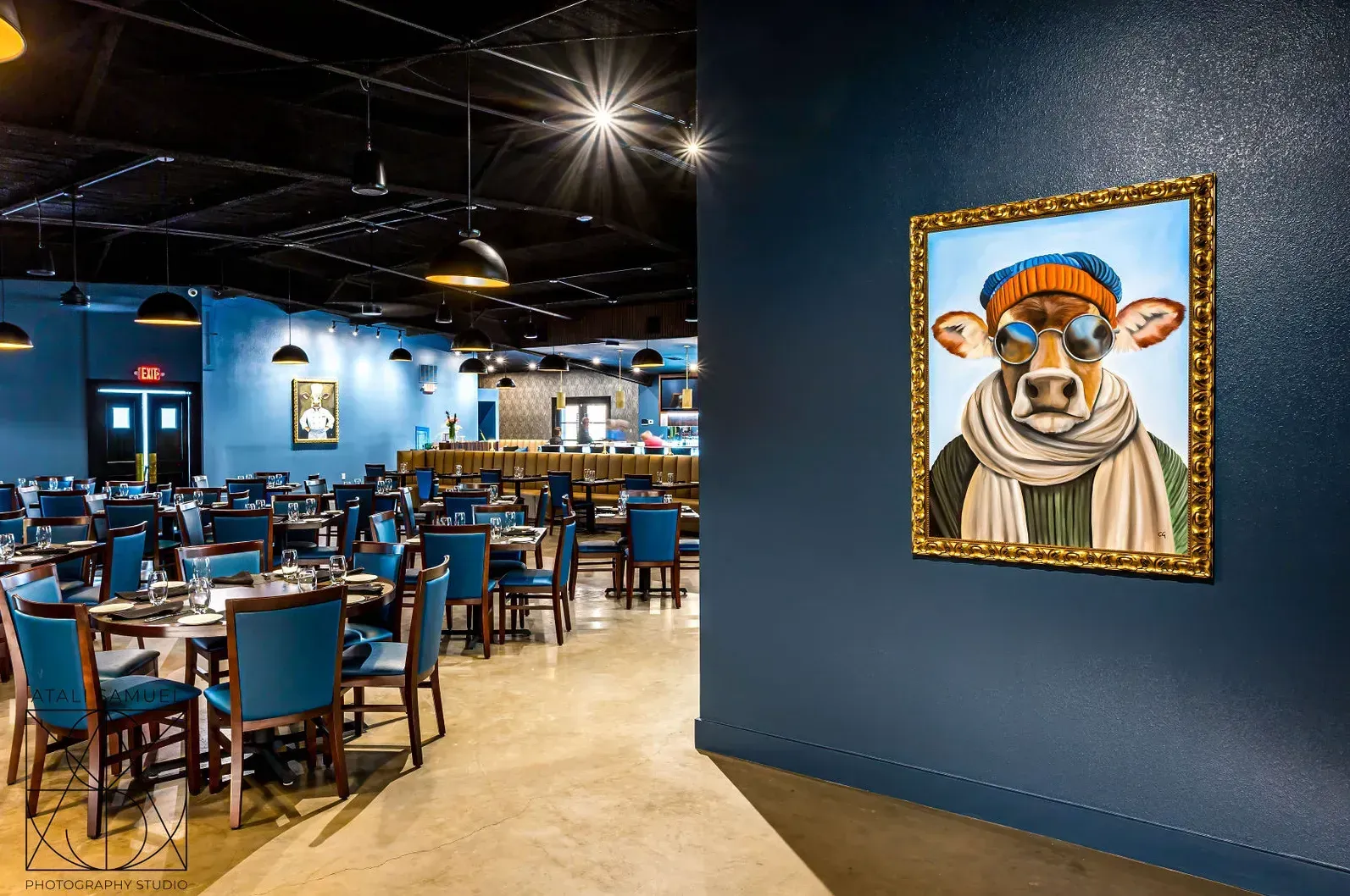 Restaurant interior with blue walls, tables, and chairs, featuring artwork of a cow wearing sunglasses.