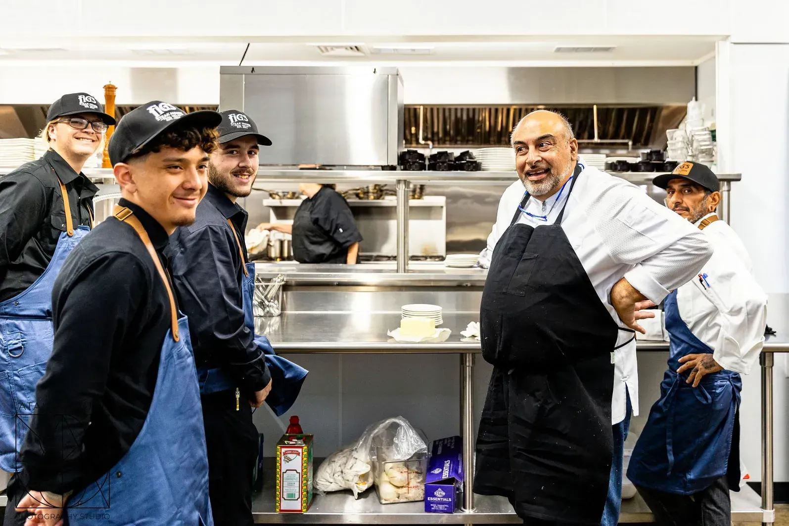 Chefs in a kitchen. Smiling chef in apron and white coat with other staff in black uniforms and aprons.