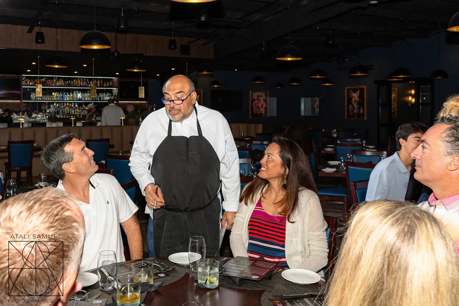 Chef in black apron talking to restaurant patrons. Dark interior, bar in background.