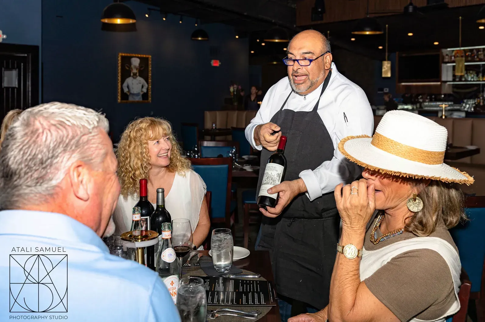 Chef presenting a wine bottle to diners at a restaurant table.