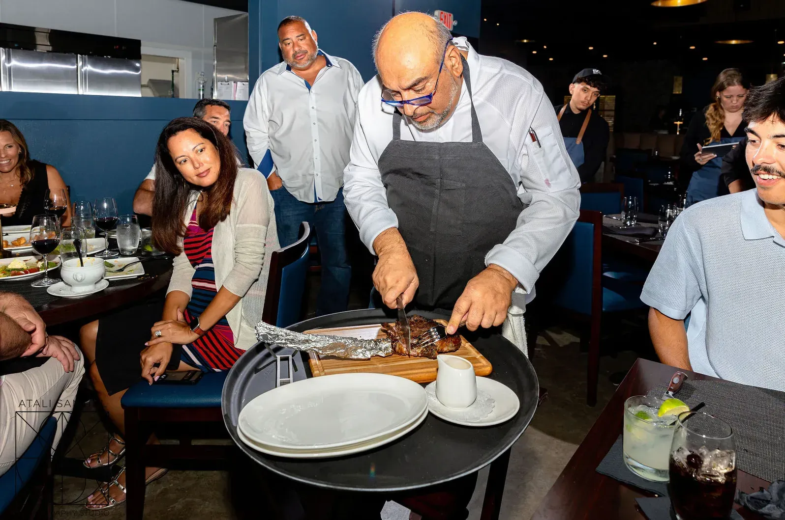 Chef carving roasted meat at a table in a restaurant. Guests watch.