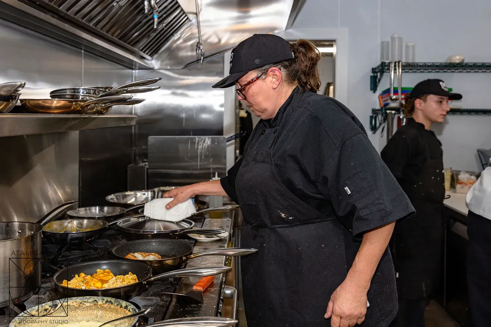 Chef in black uniform cleans a pan on a commercial stove in a restaurant kitchen.