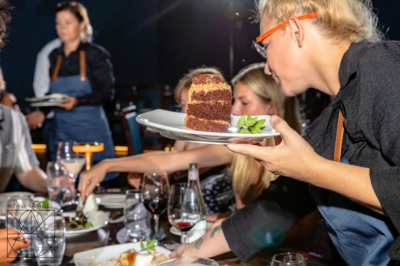 Server presents a slice of chocolate cake at an outdoor dinner table with guests reaching for it.