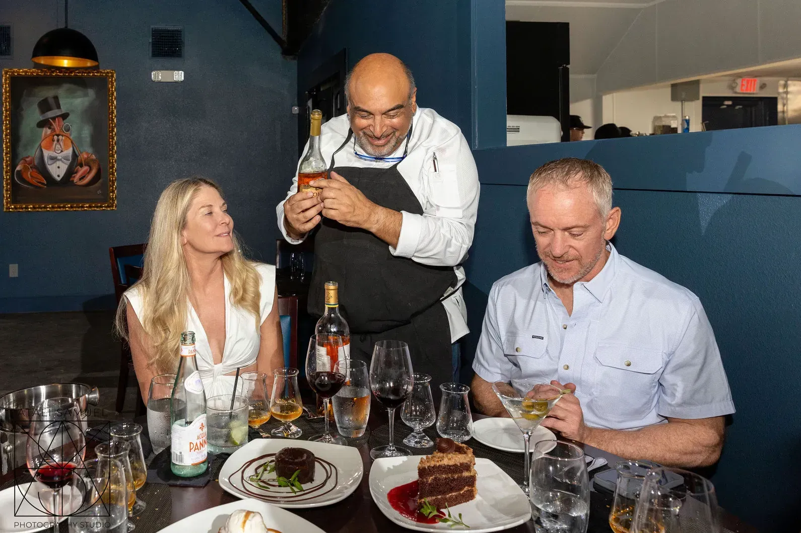 Chef presenting a drink to two diners at a restaurant table with food and wine.
