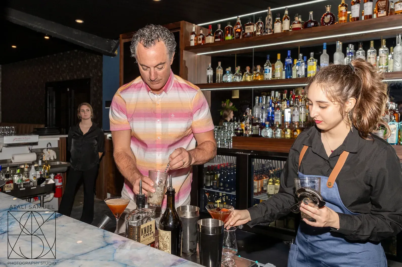 Two bartenders mixing cocktails at a bar. Bottles line shelves behind them.