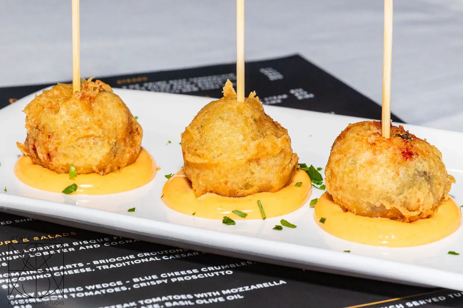 Three fried food balls on orange sauce, skewered with toothpicks, served on a white plate.