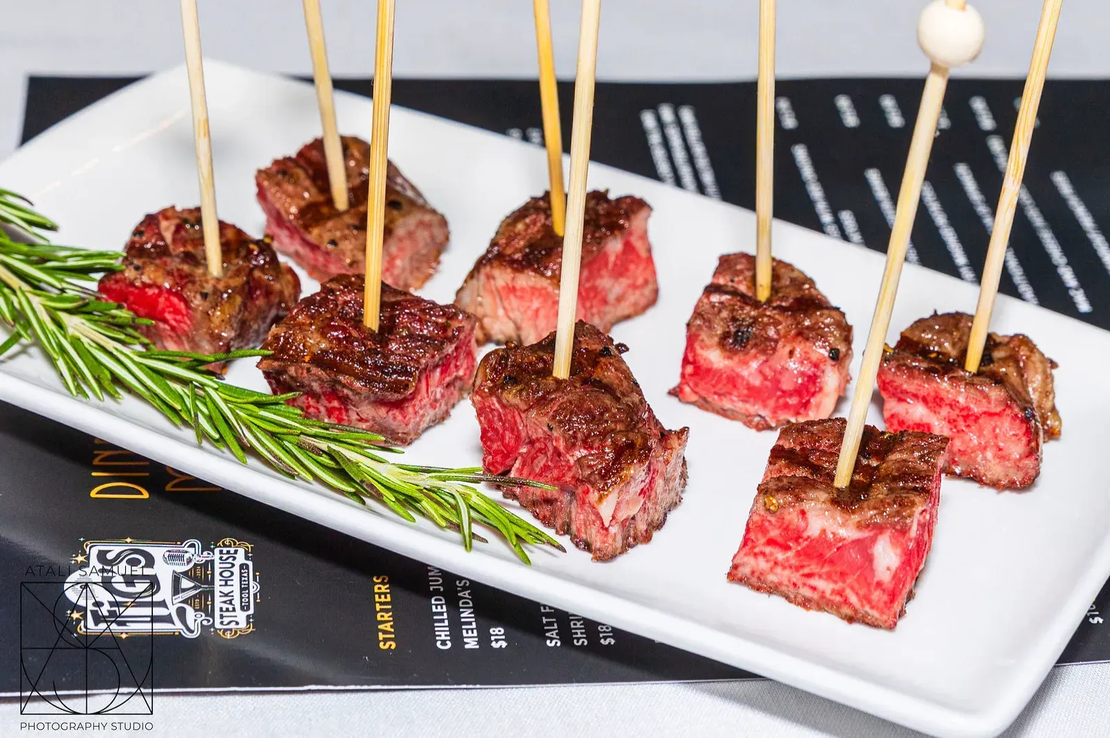 Steak bites on skewers on a white plate with rosemary sprigs.