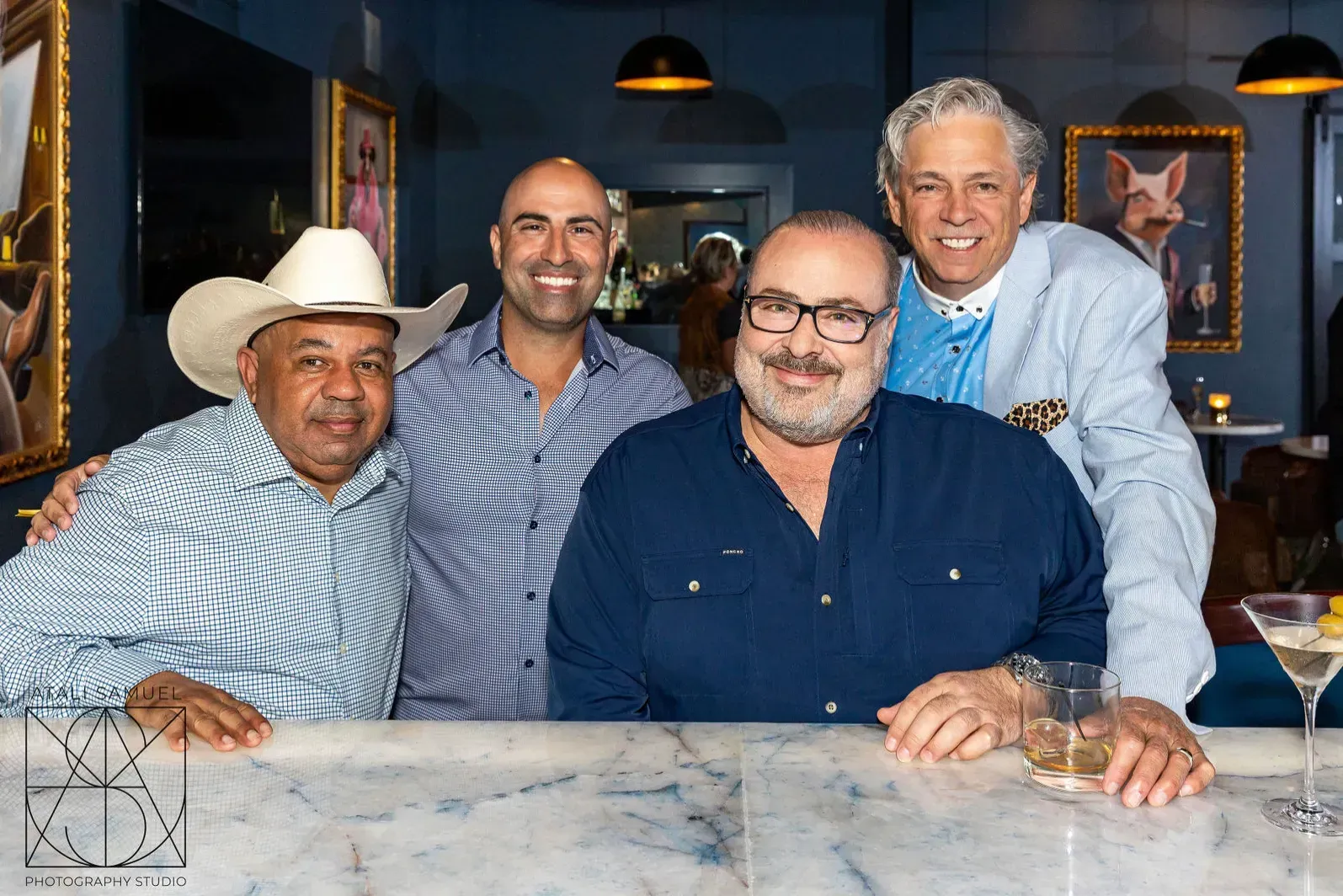 Four men at a bar, smiling. One in a cowboy hat, another in a blue shirt, the central figure in a button-up, and the fourth in a blazer.