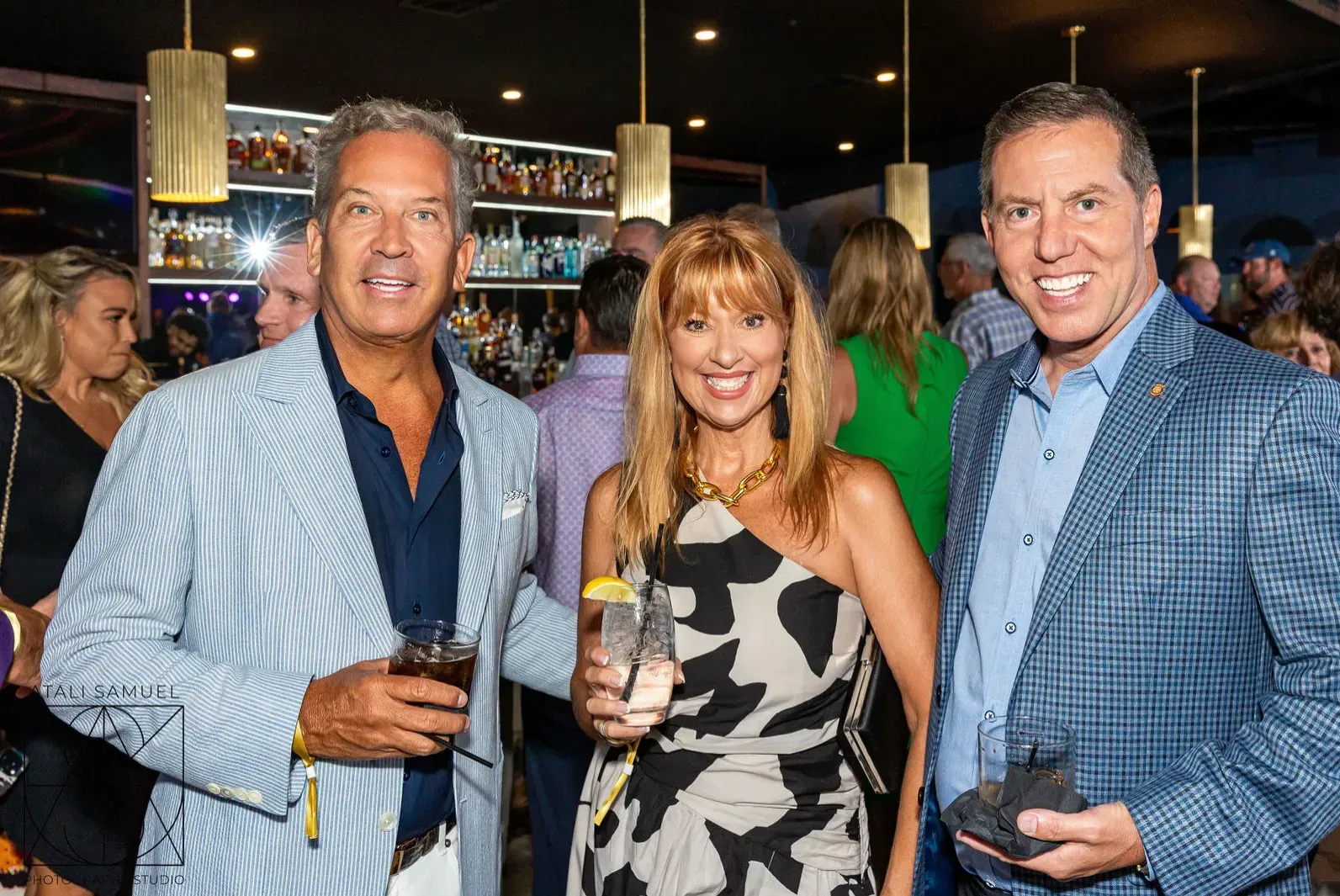 Three people smiling at an event; bar background, one wearing blue striped jacket, holding a drink.