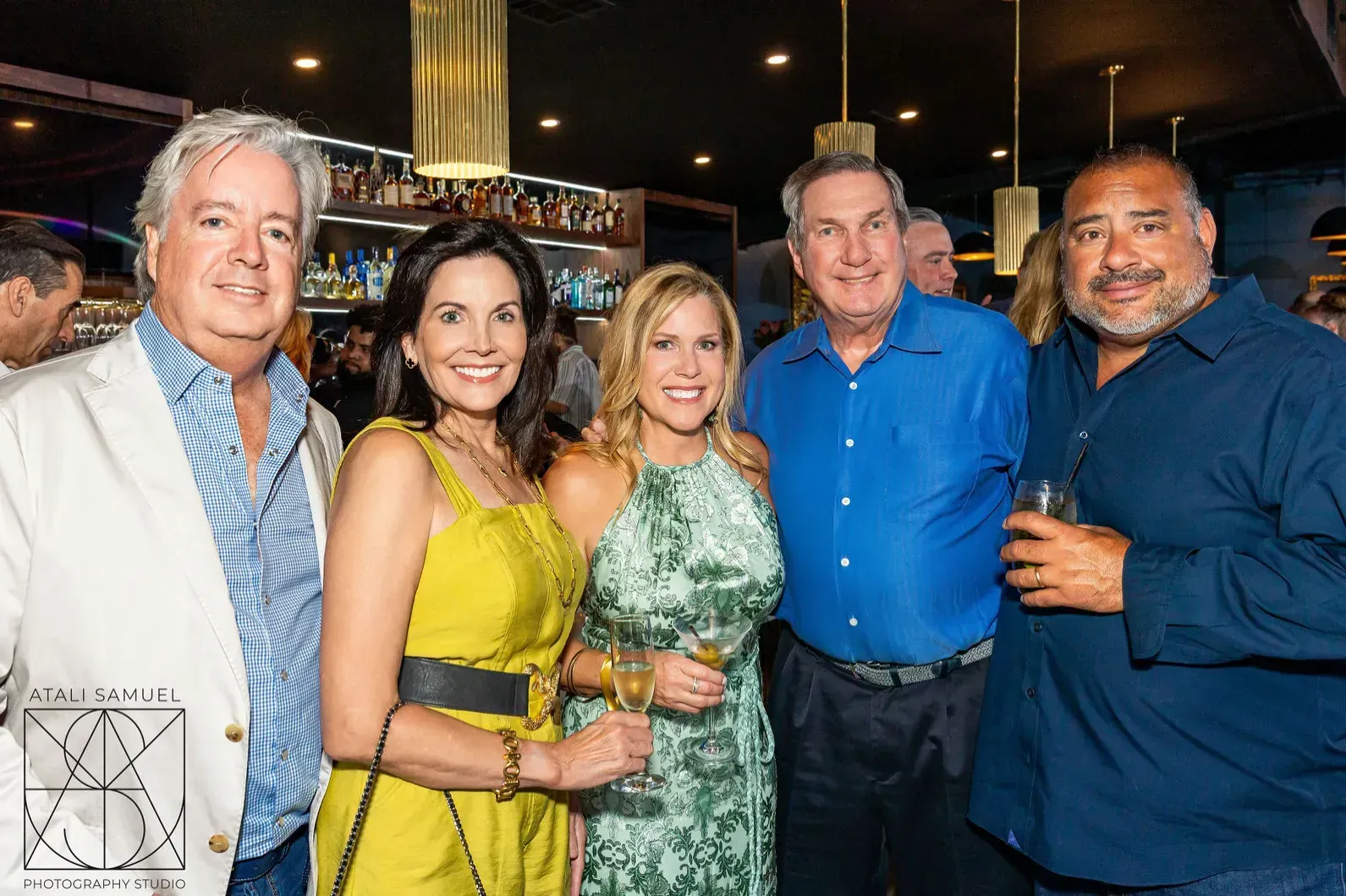 Five people smiling in a bar. Three women wear dresses, the men button-down shirts. Bright setting with bar in background.