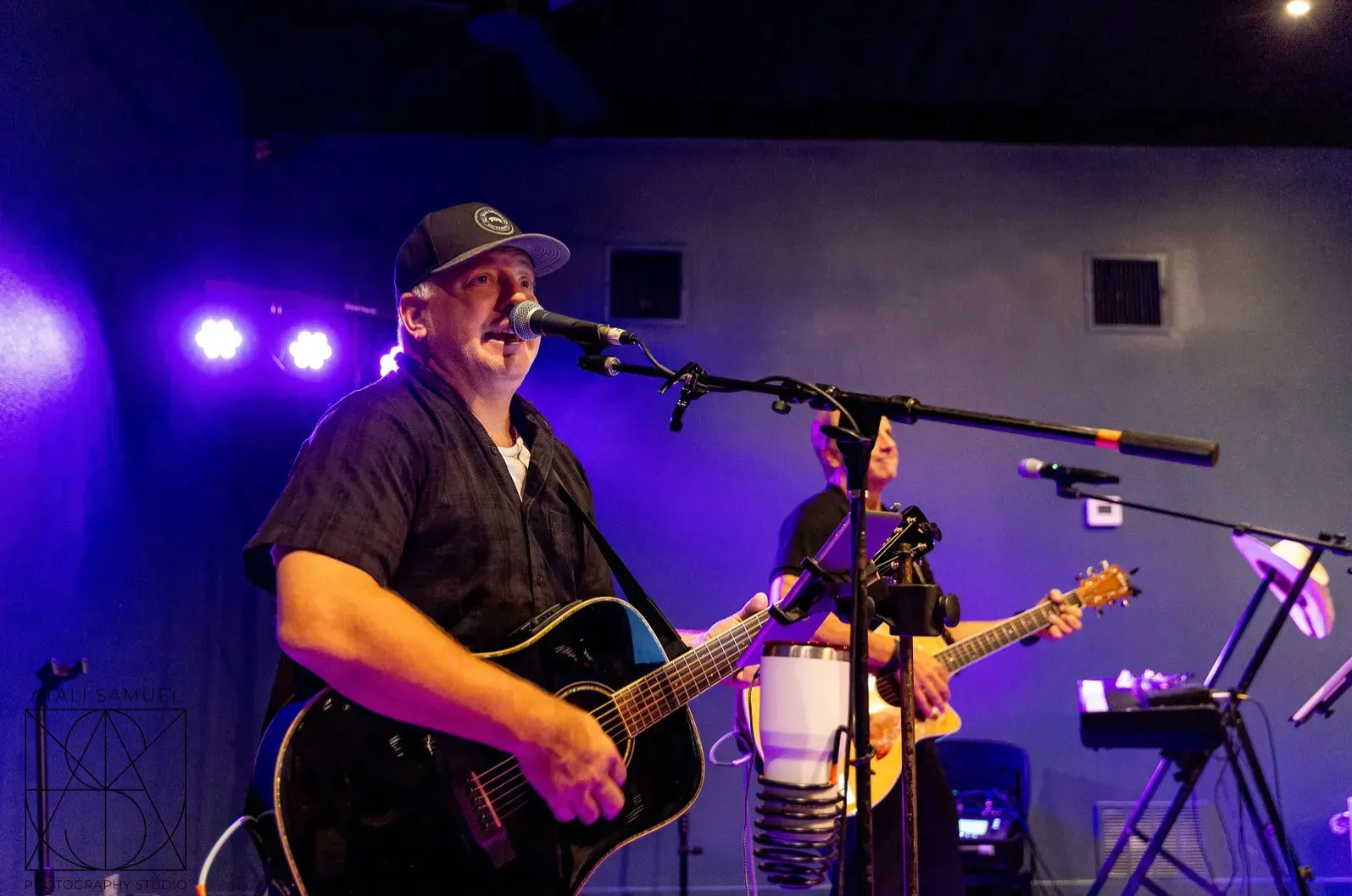 Two musicians performing on stage: one singing and playing guitar, the other playing guitar near a drum. Purple lighting.