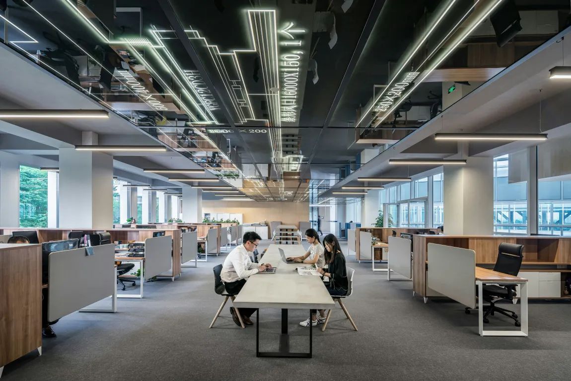 an office with a long table in the middle and people sitting across from each other on laptops