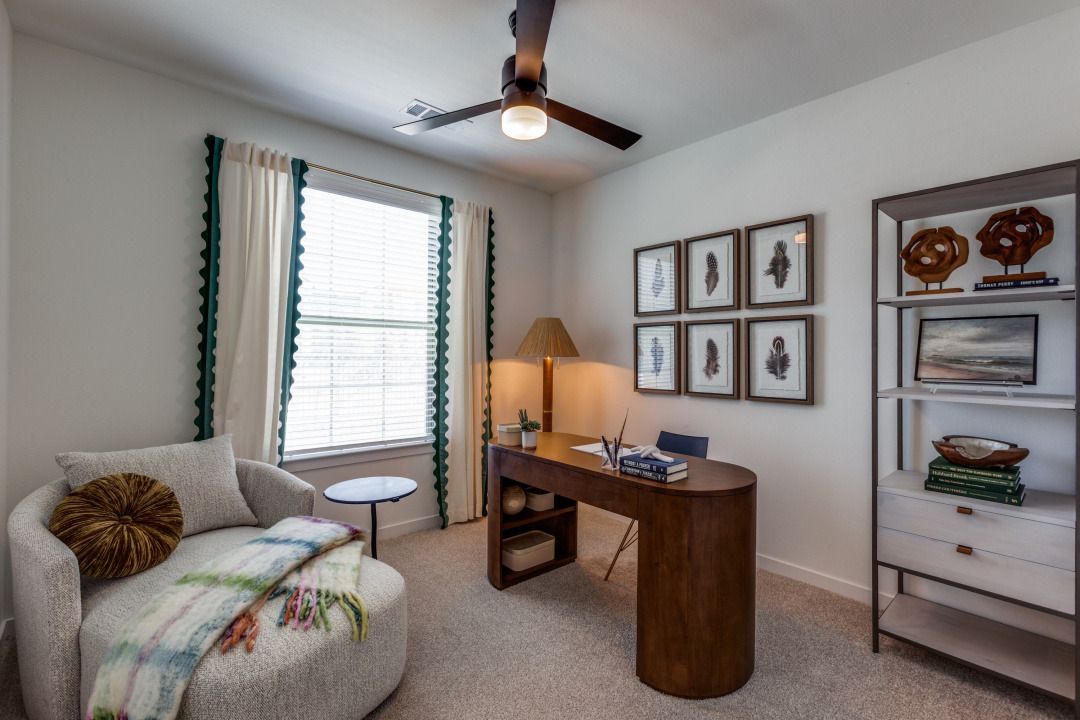 A bedroom with a desk , chair , ottoman and ceiling fan.