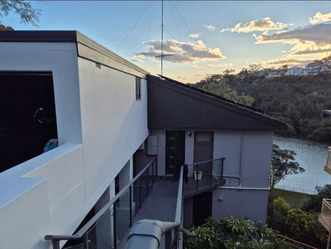 Person in protective suit sprays paint on a corrugated metal roof on a sunny day, with mountains in the background.