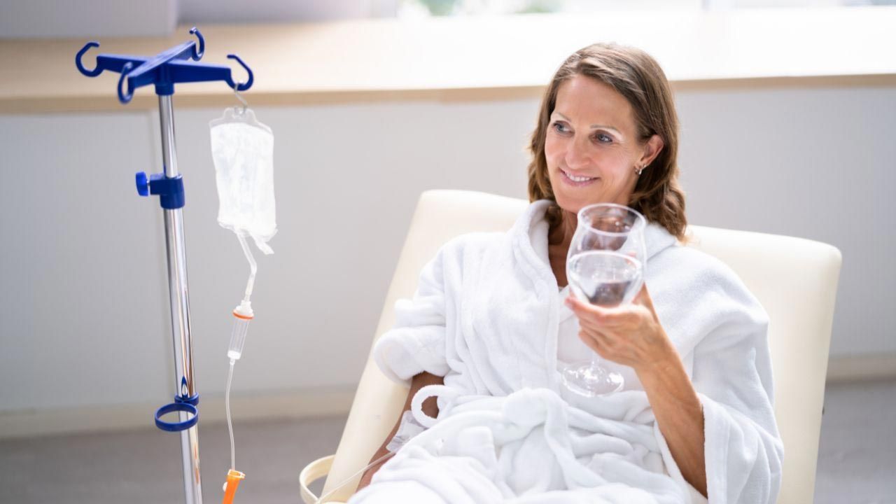 A woman is sitting in a hospital chair holding a glass of water. - IV Nutrition