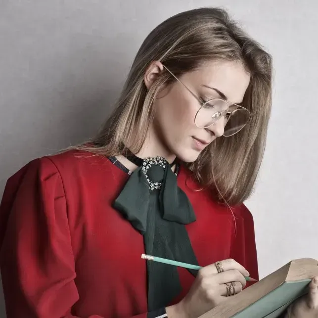 Woman in red blouse with a green bow writing in a book, wearing glasses.