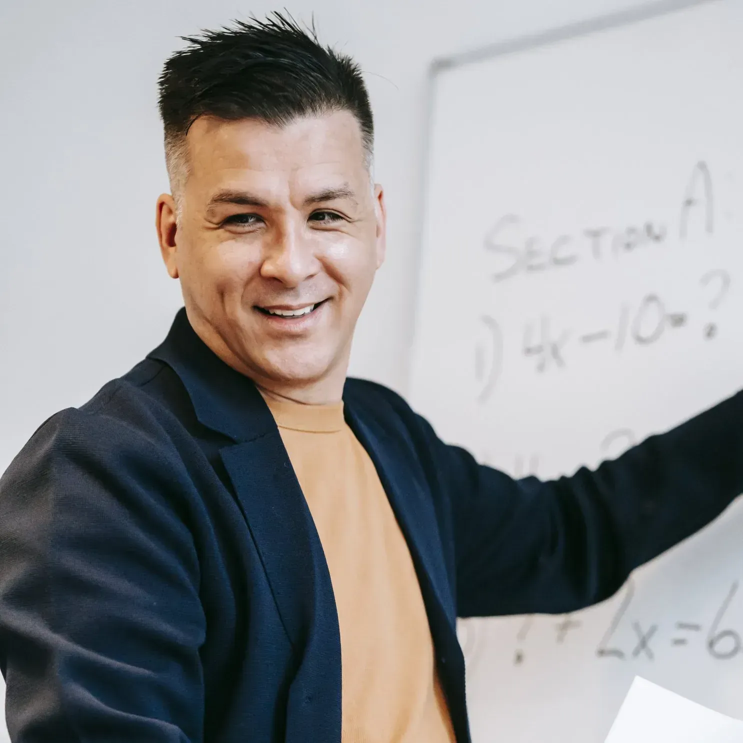 Man smiles, points to a whiteboard with math problems. He wears a blazer over a yellow shirt.