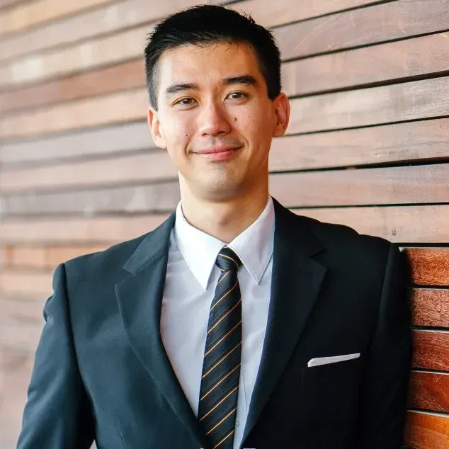 Man in suit, smiling, leans against a wooden wall, wearing a striped tie.