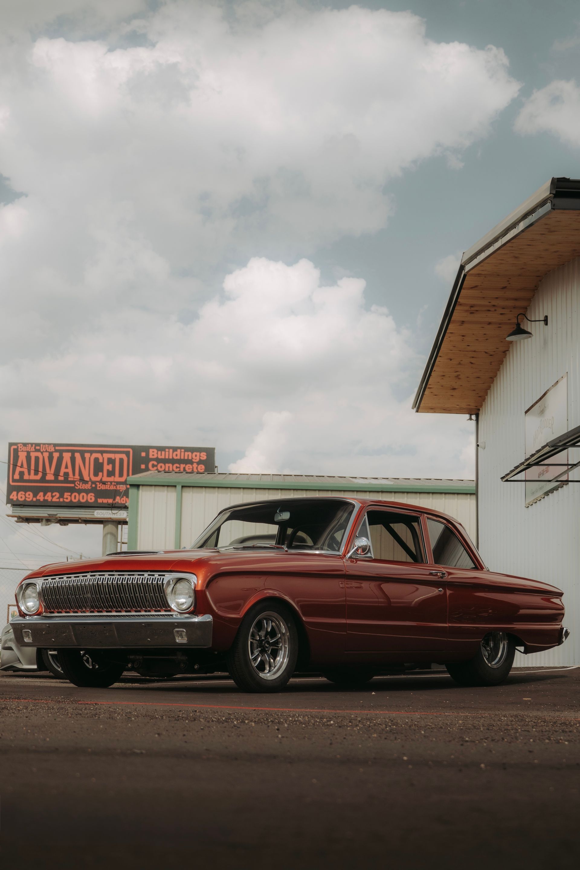 Red classic Ford Falcon drag racer in front of a white building with ADVANCE signs under a cloudy sky.