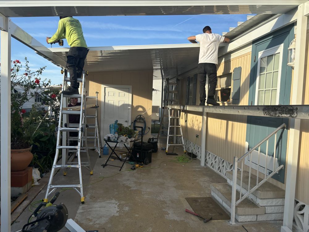 Two workers building a white patio cover attached to a light blue and tan house.