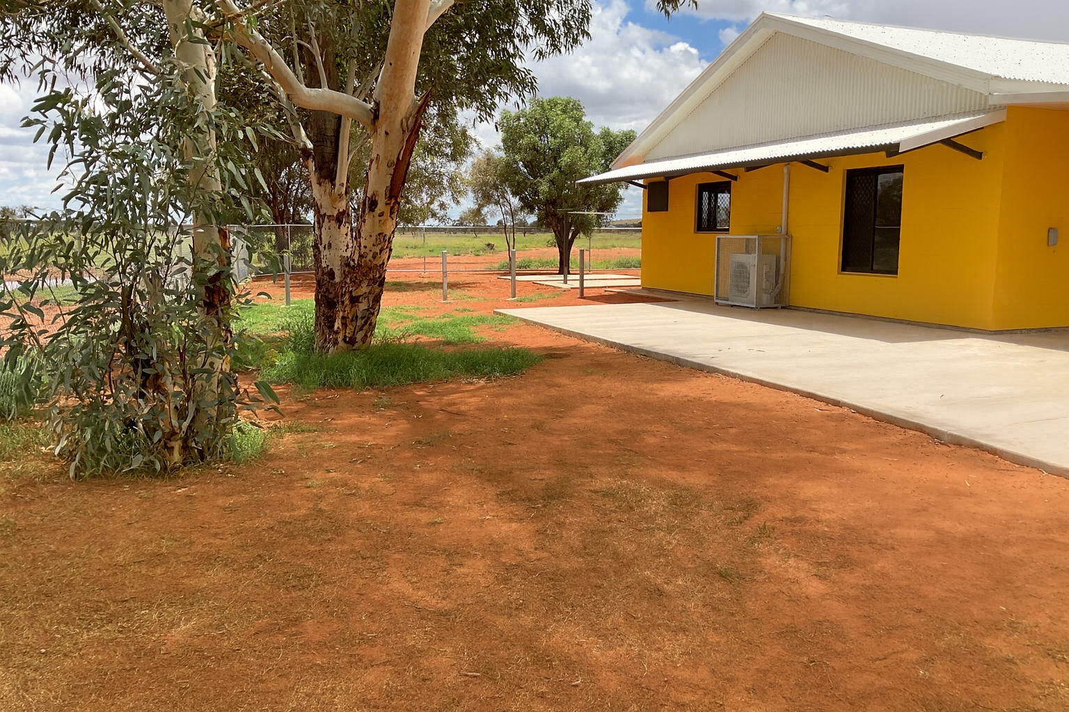 Yellow building with white roof, concrete path, red dirt, and trees under a partly cloudy sky — Pedersen NT Pty Ltd in Ciccone, NT