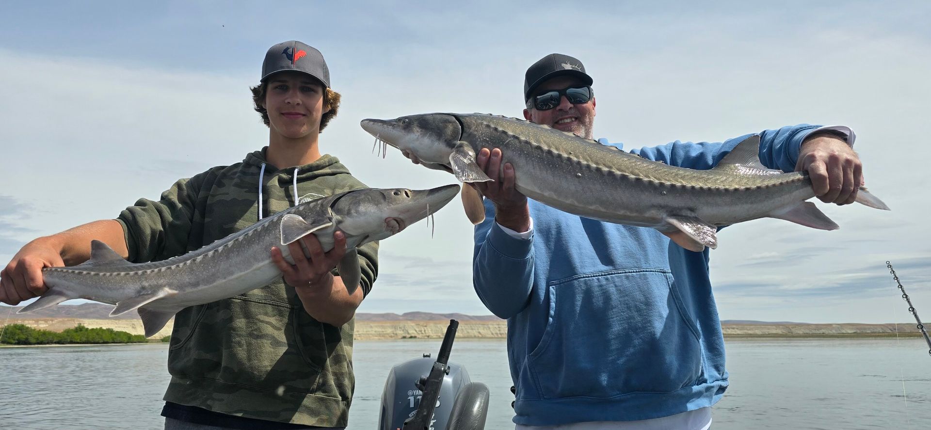 Two people on a boat hold up large fish, likely sturgeon, in front of a scenic outdoor backdrop.