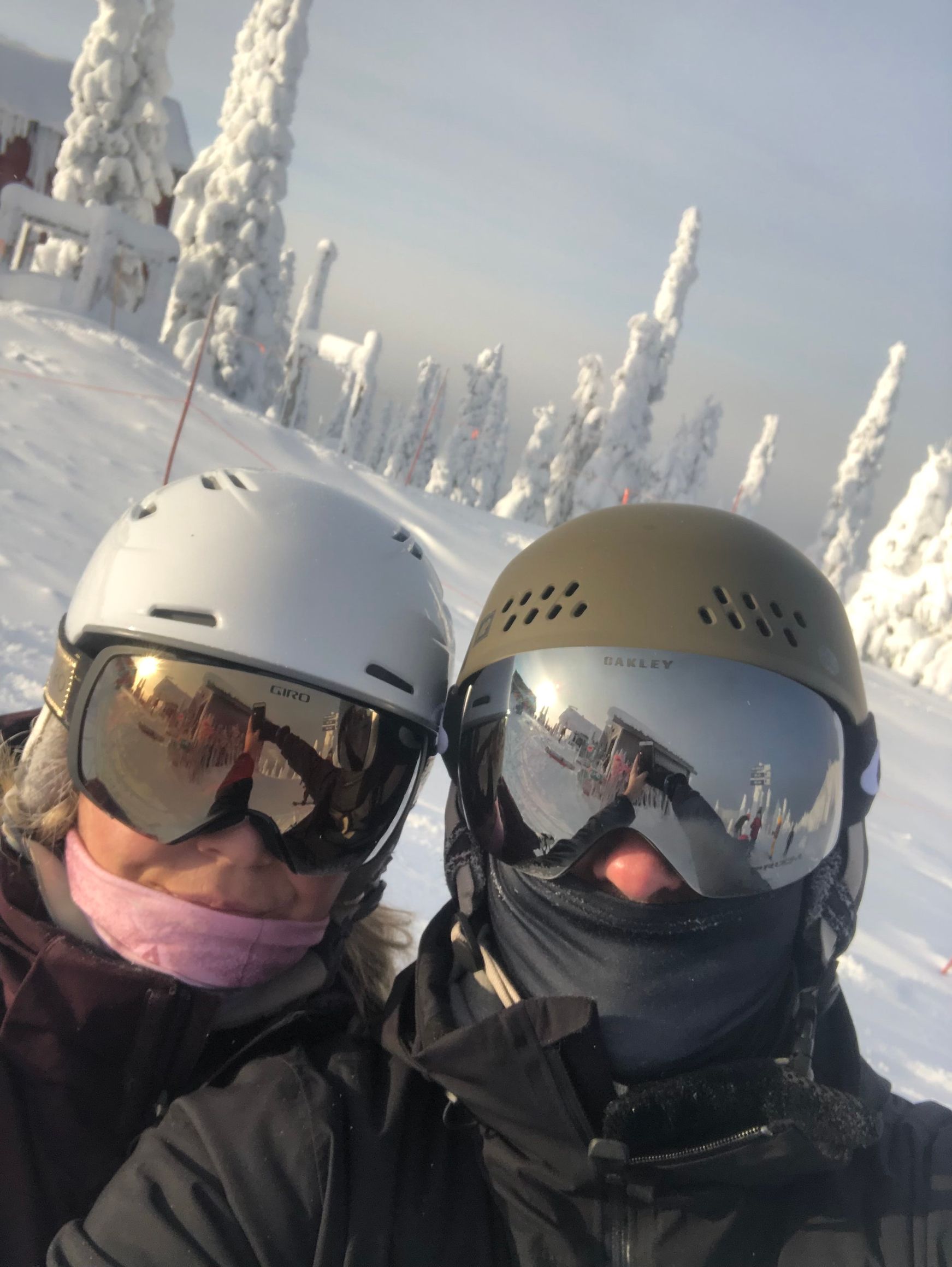 Two people in ski helmets and goggles, on a snowy slope, with frosted trees.