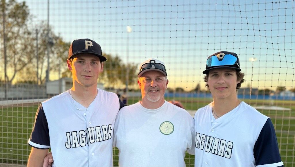 Three people in baseball uniforms: two young men flanking an older man, all smiling, on a baseball field.