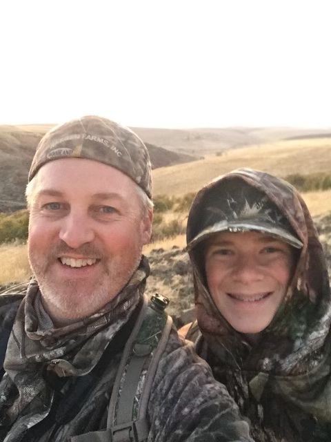 Man and boy in camouflage gear smiling outdoors with a hilly landscape in the background.