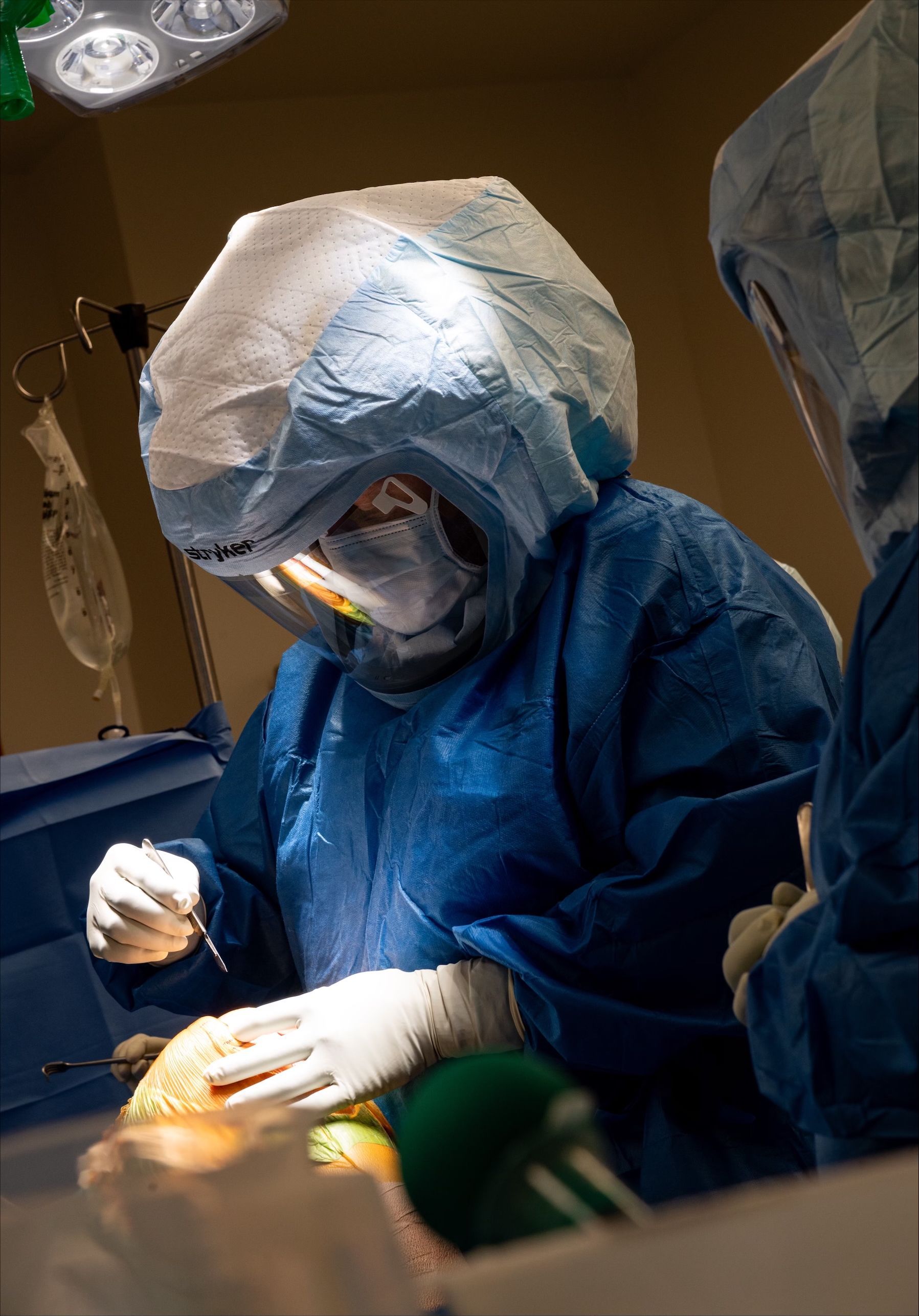 Surgeon in blue gown and hood operates in a sterile surgical room, using tools on a patient.