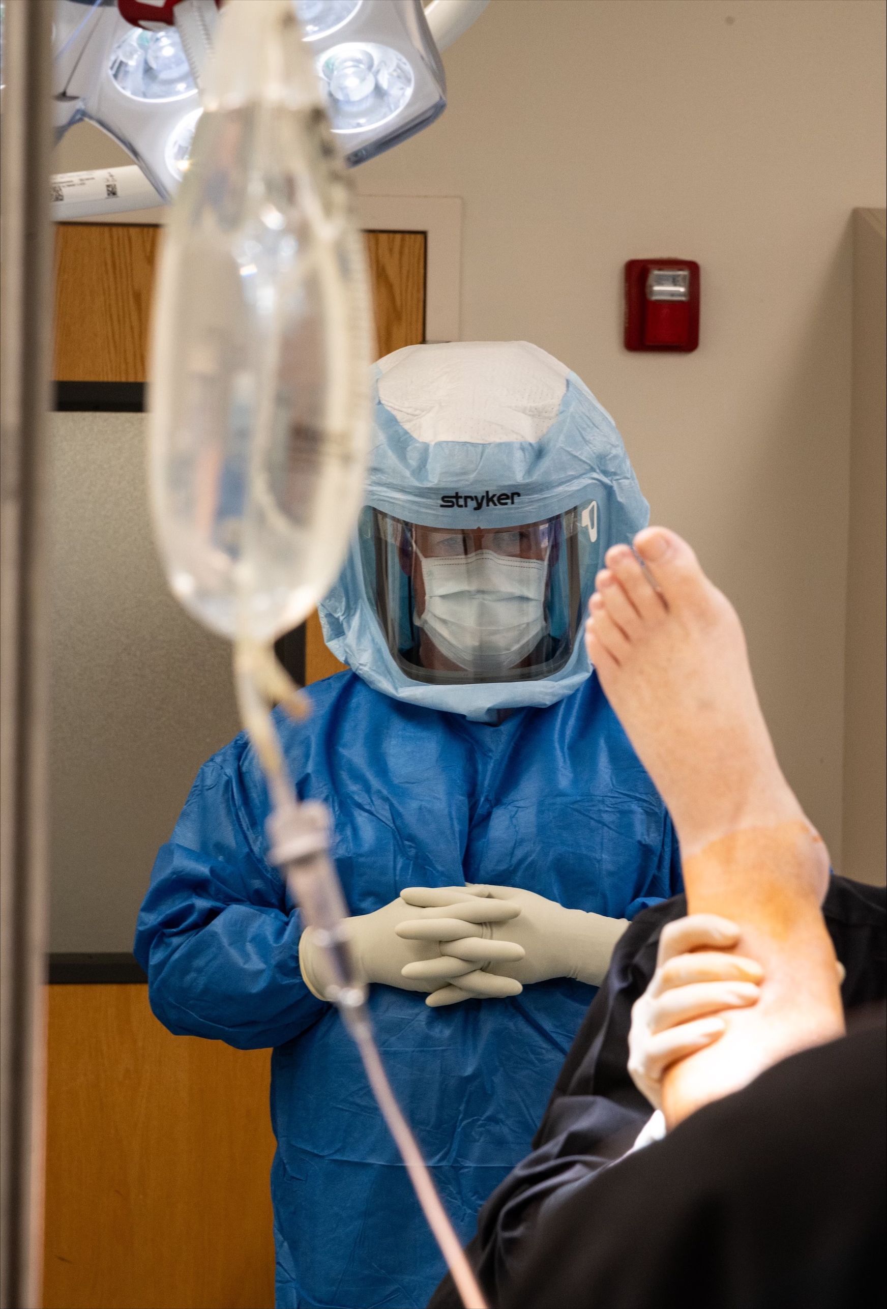 Surgeon in protective gear observing a patient's foot in operating room; IV bag in the foreground.