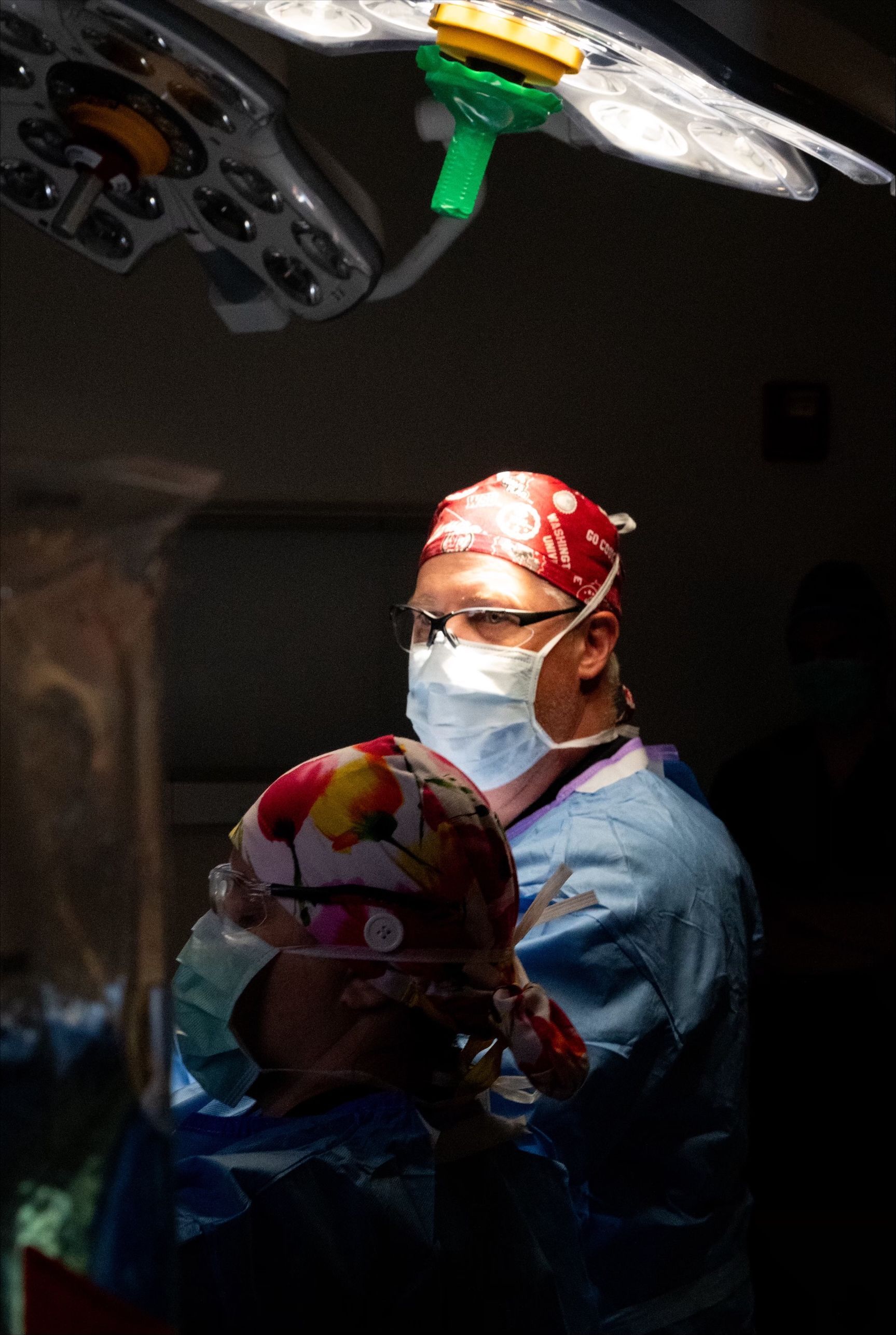 Surgeon in operating room, wearing mask, holding a head cap, with surgical lights overhead.