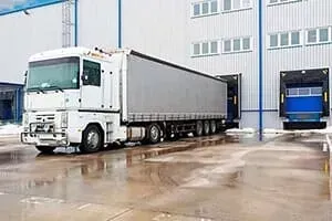 White semi-truck at a loading dock of a warehouse, gray trailer, blue doors, light exterior.