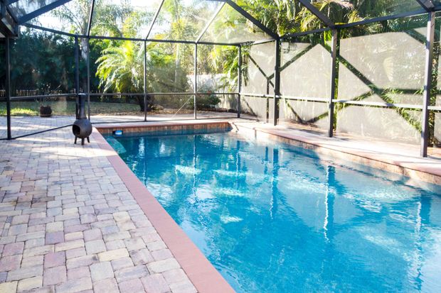 A swimming pool with blue water enclosed by a screened lanai, surrounded by light-colored pavers. A swimming pool with blue water enclosed by a screened lanai, surrounded by light-colored pavers.