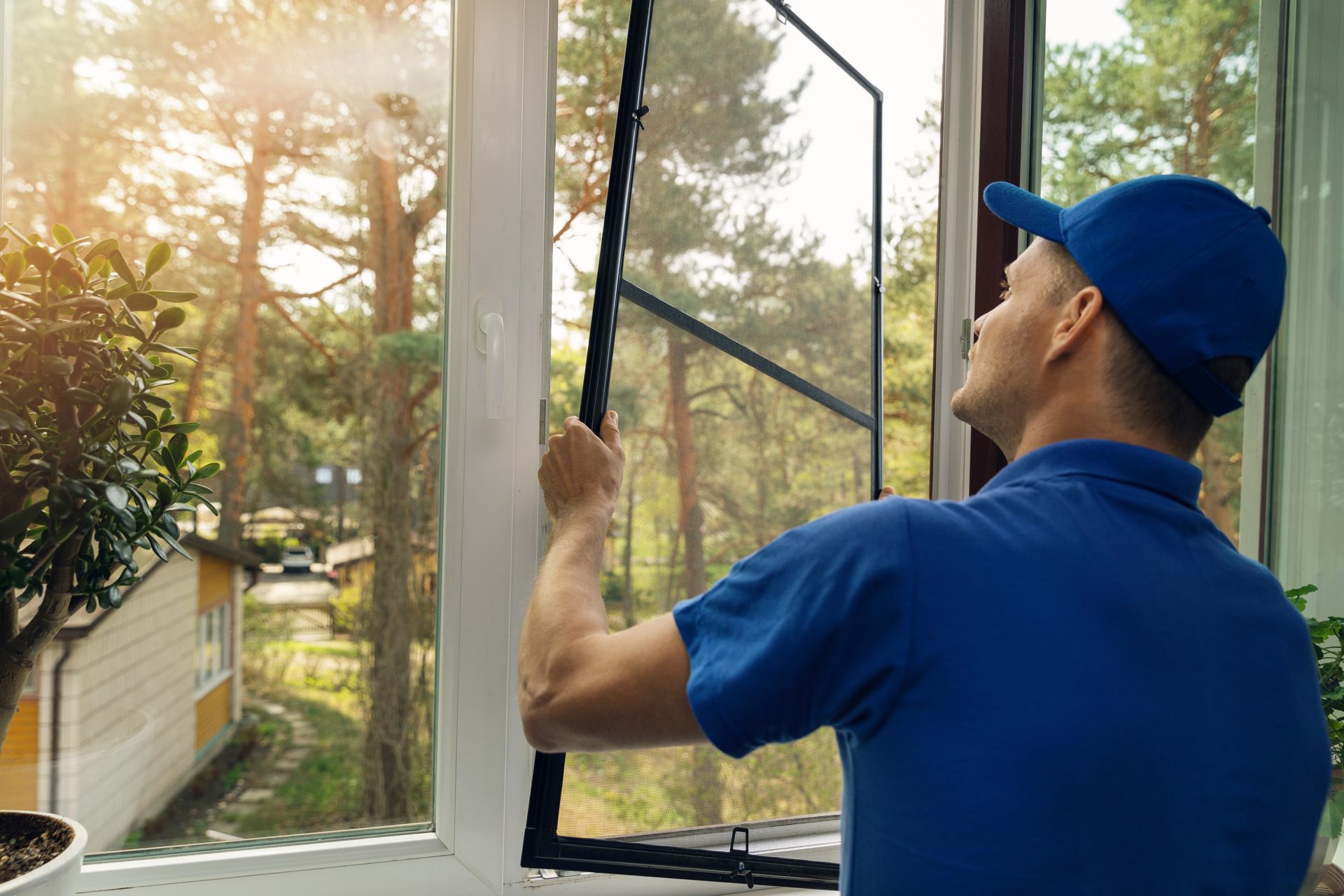 A worker in a blue uniform installs a window screen, with a view of a garden and trees visible through the window.