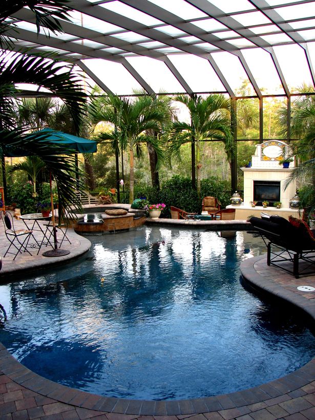 Indoor swimming pool with a fireplace, patio furniture, and palm trees under a glass-enclosed structure. Indoor swimming pool with a fireplace, patio furniture, and palm trees under a glass-enclosed structure.
