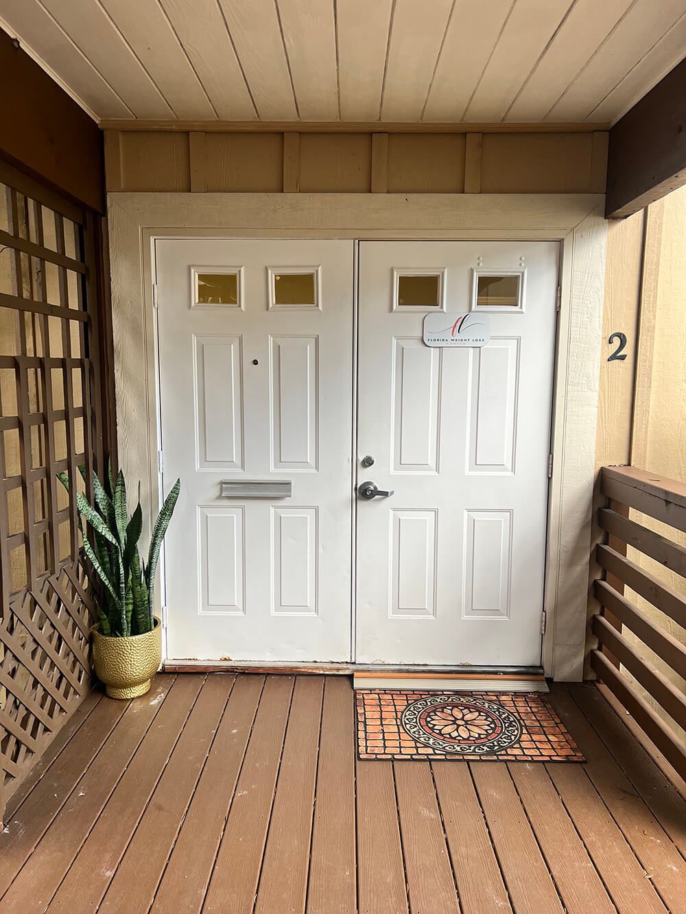 A pair of white double doors on a wooden porch, with a potted plant to the left and a decorative mat in front.