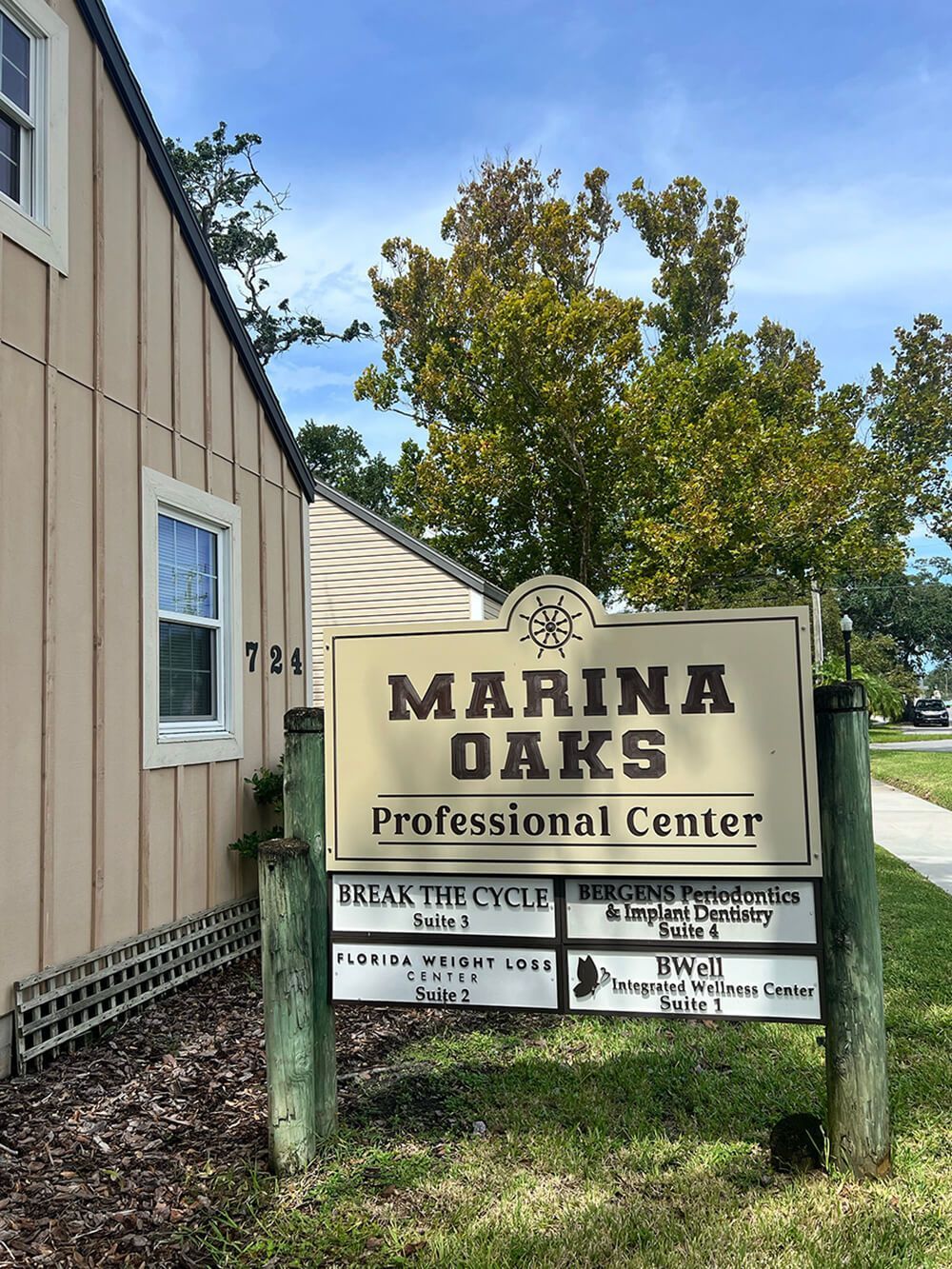A beige, outdoor sign for Marina Oaks Professional Center, listing several businesses, sits in front of a beige building.