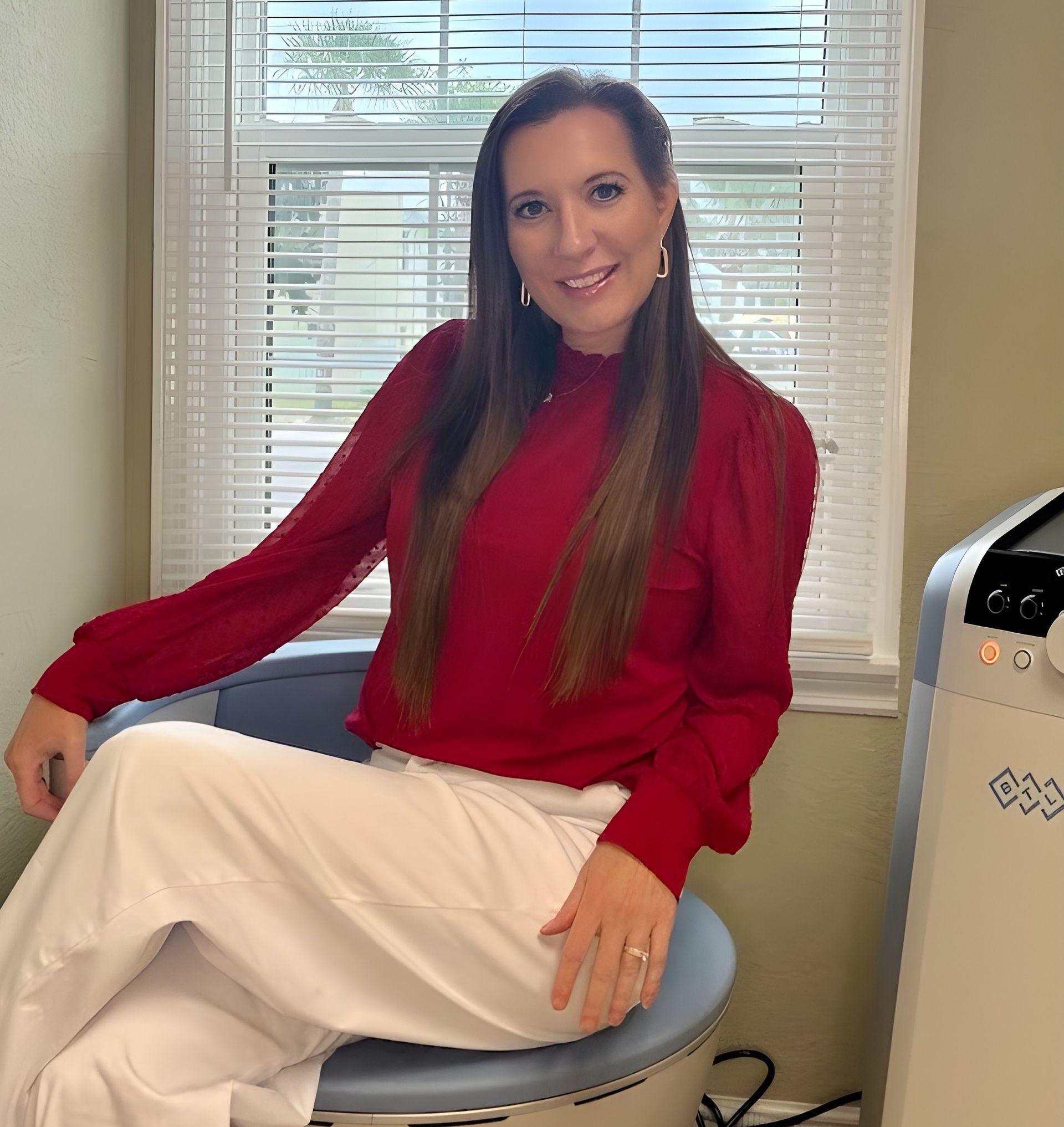 A woman with long brown hair wearing a red blouse and white pants sits in a chair in a room with blinds on the window.
