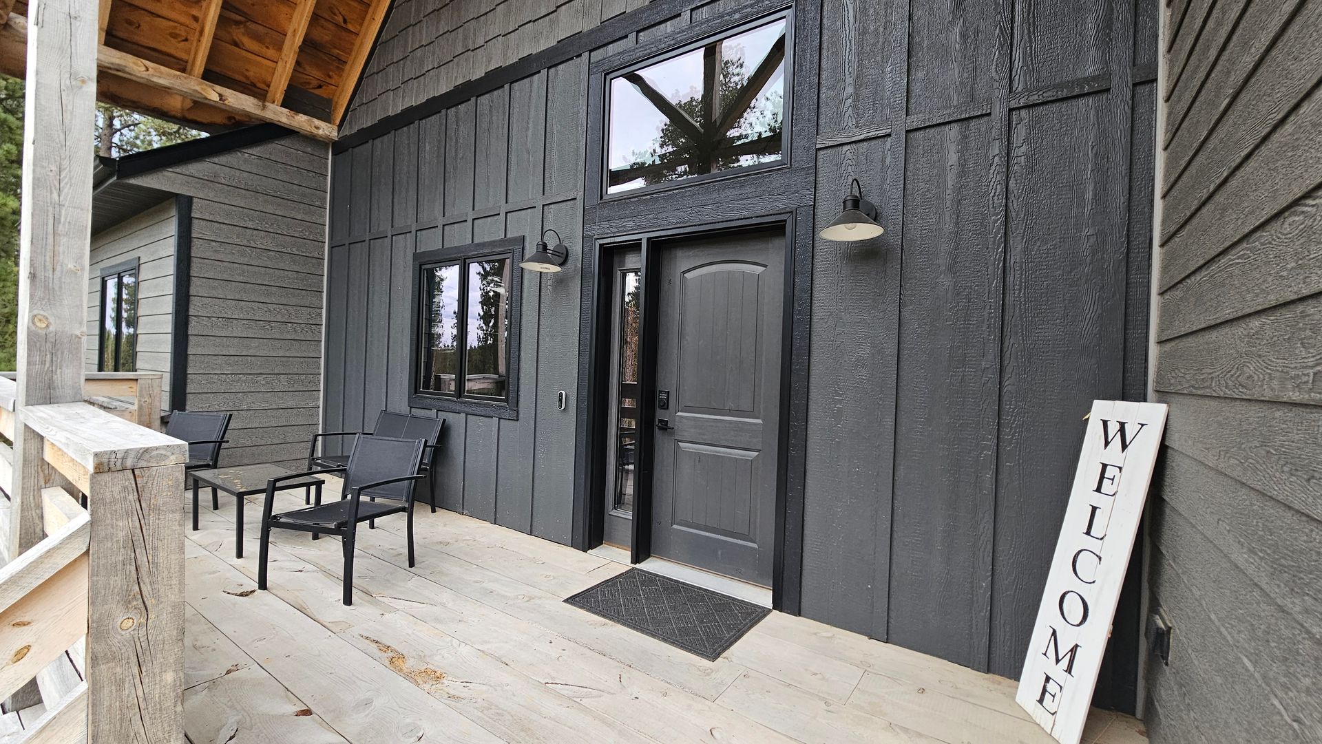 Dark gray cabin entrance with small patio, chairs, and welcome sign.