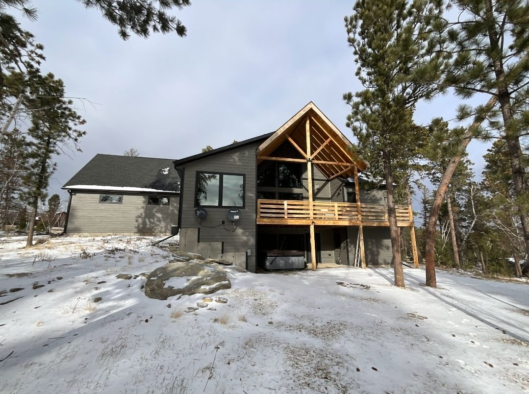 A gray cabin with a wooden deck in a snowy, wooded area.