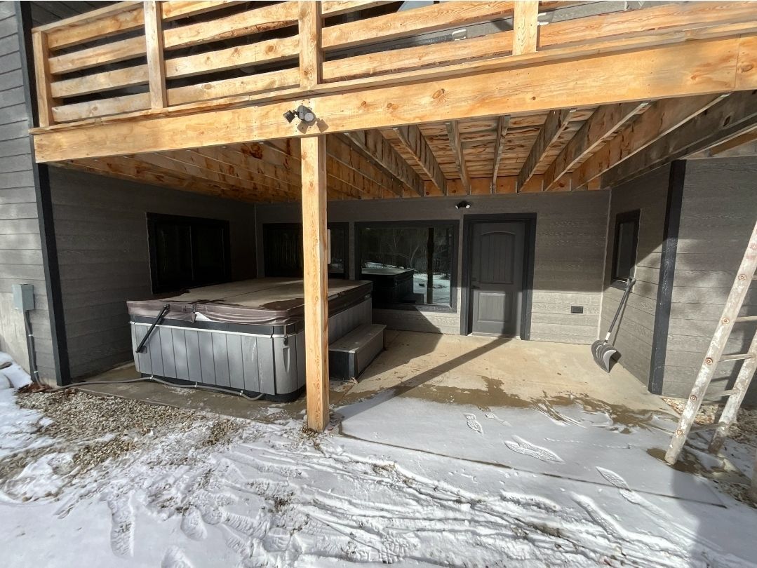 A hot tub under a wooden deck, surrounded by snow and a gray building.