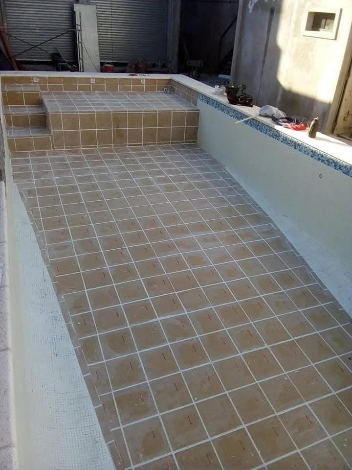 Interior de piscina vacía con suelo de baldosas y escalones. Azulejos beige, lechada blanca. Exterior soleado.