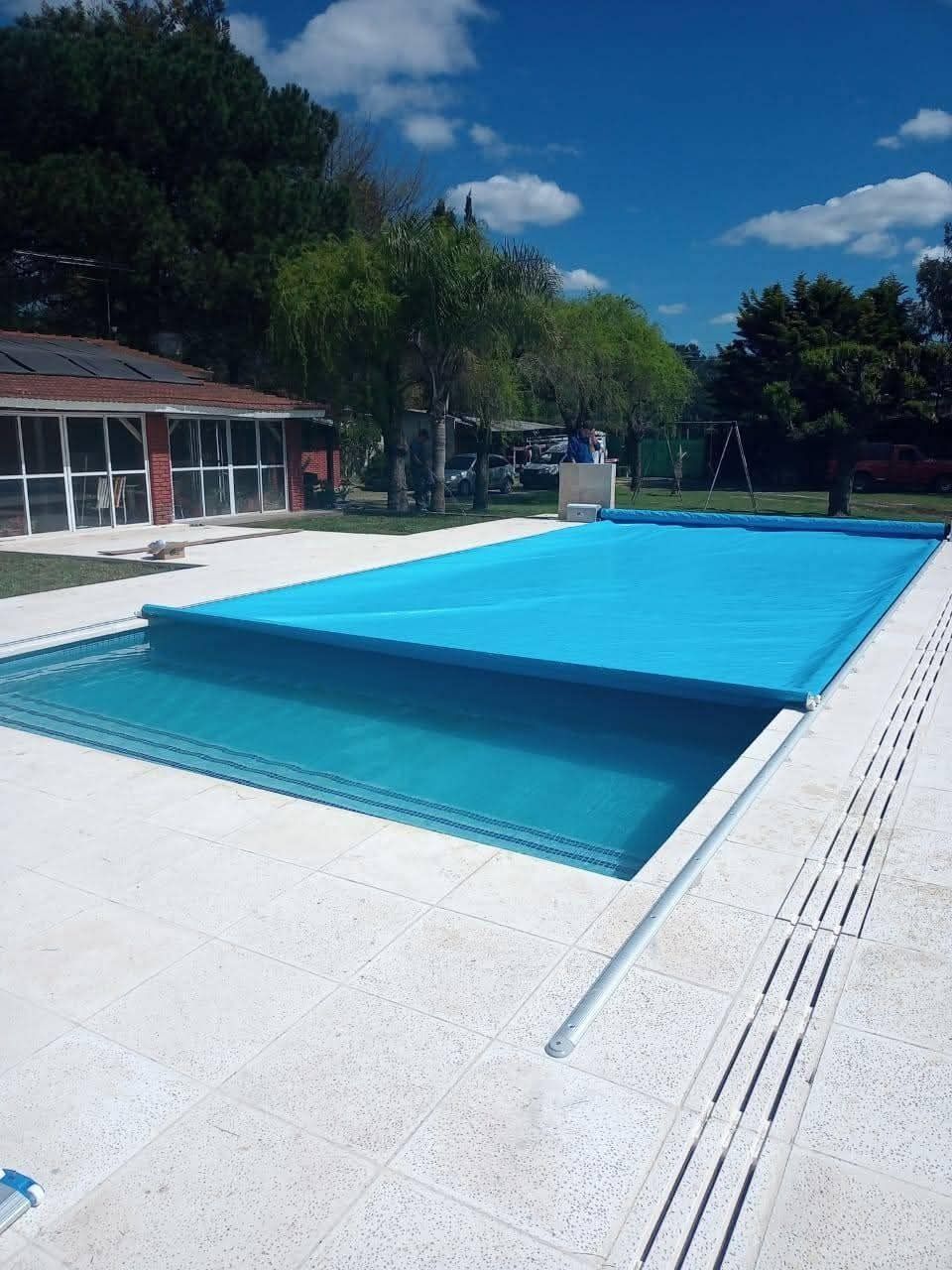 Cubierta de piscina azul sobre una piscina rectangular en un entorno exterior soleado con un edificio y árboles.