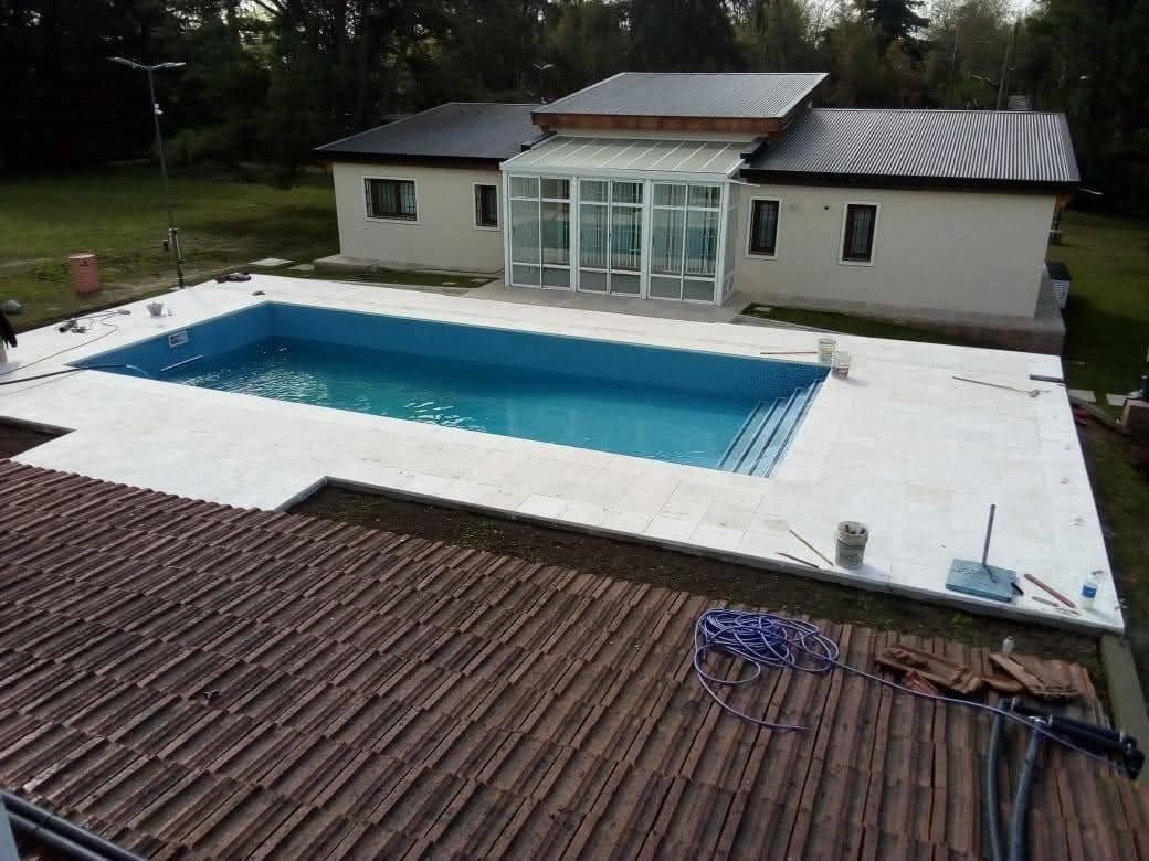 Vista de una casa con piscina, cerramiento de cristal y techo de tejas marrones. Una terraza blanca rodea la piscina azul.