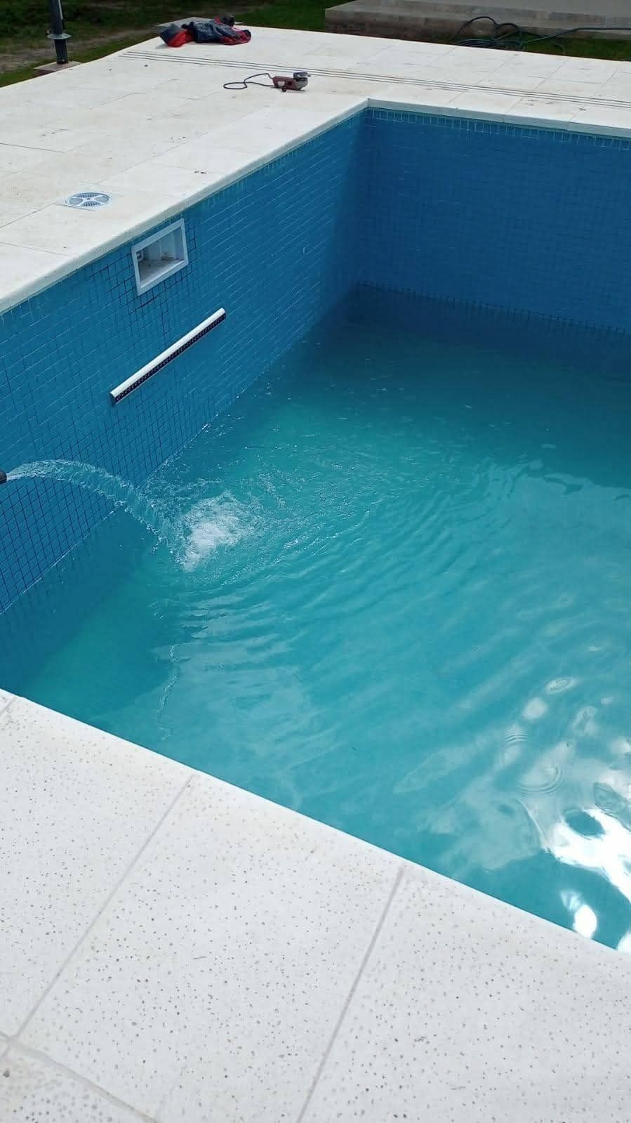 Piscina rectangular terminada, agua turquesa, borde de baldosas blancas, interior de mosaico azul.