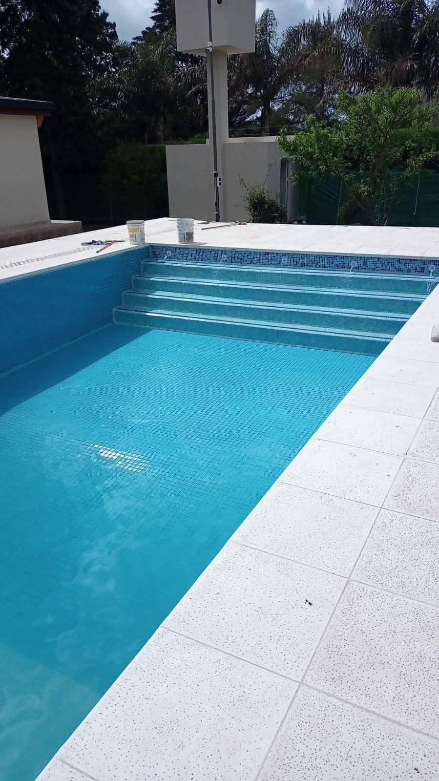 Una piscina azul con escalones, rodeada de piedra blanca y un pequeño edificio blanco al fondo.