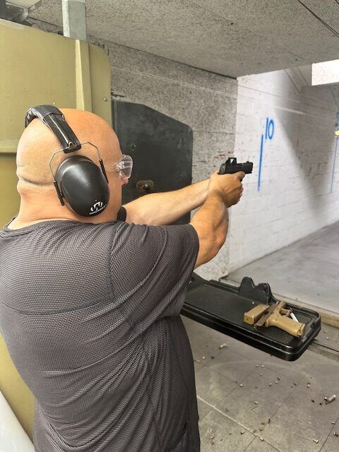 Man at shooting range, wearing ear and eye protection, aiming a handgun at a target. A second gun rests in a tray nearby.