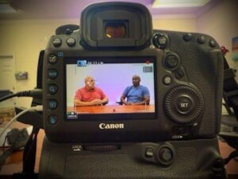 Back of a Canon camera filming two men seated at a table; screen shows them talking in front of a light purple backdrop.